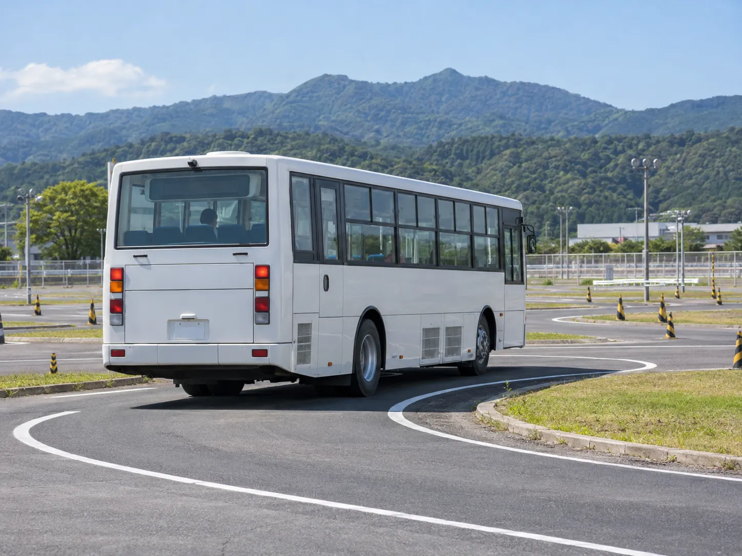 教習所のコースを走る白い大型バスを後ろから見たところ。奥に山並み